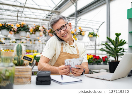 A Caucasian florist, happy with money in hand, smiles while on a phone call in his shop, surrounded by plants and flowers. 127119220
