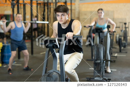 Dedicated active strong young man doing cardio workout on stationary bicycle as hard cross-training in gym indoors 127120343