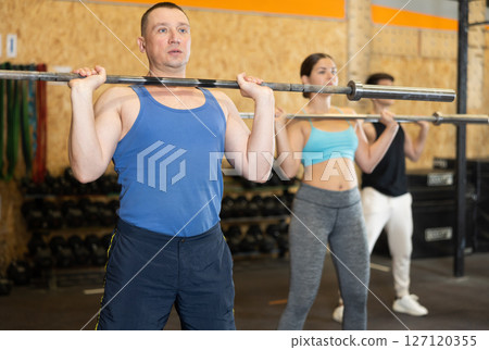 Focused middle-aged man in activewear exercising with barbell during group workout in gym health center. CrossFit healthy concept Focused middle-aged man in activewear exercising with barbell during group workout in gym health center. CrossFit healthy concept 127120355