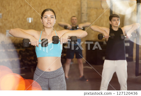 Sportive strong young female in activewear lifting dumbbells during group workout class in gym indoors. Functional training concept 127120436