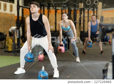 Motivated young man practicing exercises with kettlebell near other people in gym 127120468