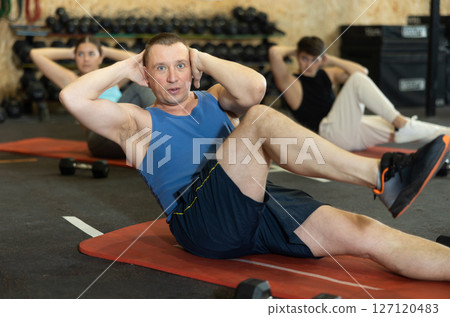 Concentrated middle-aged man doing abdominal exercise on yoga mat during group workout in gym. CrossFit healthy concept 127120483