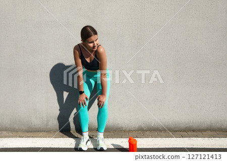 Athletic woman resting after workout against a wall, wearing stylish sportswear, focused on hydration with a water bottle, fitness lifestyle concept Athletic woman resting after workout against a wall, wearing stylish sportswear, focused on hydration with a water bottle, fitness lifestyle concept 127121413