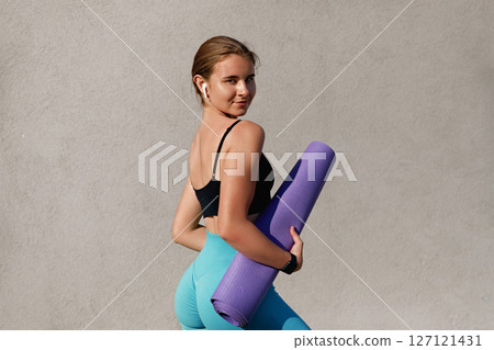 Young woman in activewear holding a yoga mat, showcasing fitness lifestyle, with a confident pose against a textured gray wall, embodying health and wellness in a vibrant atmosphere 127121431