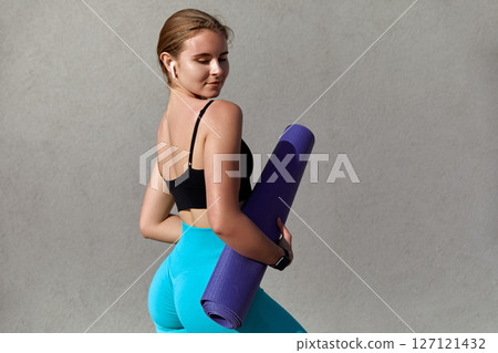 Young woman in black sports bra and turquoise leggings holding a purple yoga mat, posing confidently against a textured gray background, embodying fitness and wellness lifestyle concepts 127121432