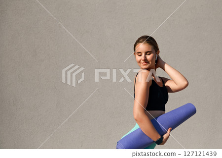 Young woman in activewear holding a yoga mat, smiling and enjoying a moment of relaxation, with a neutral background that enhances her joyful expression and promotes a healthy lifestyle concept 127121439