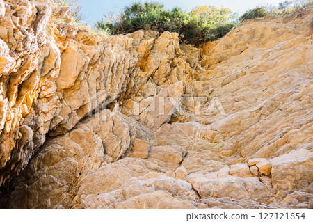 Sunlit rock wall with jagged natural lines and weathered surface. Geological texture, sediment layers, and erosion in coastal terrain. Sunlit rock wall with jagged natural lines and weathered surface. Geological texture, sediment layers, and erosion in coastal terrain. 127121854