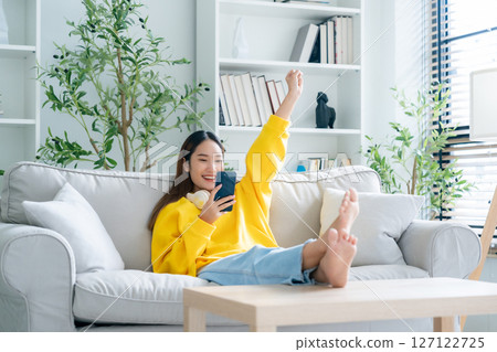 Happy young asian woman relax on comfortable couch at home texting messaging on smartphone 127122725