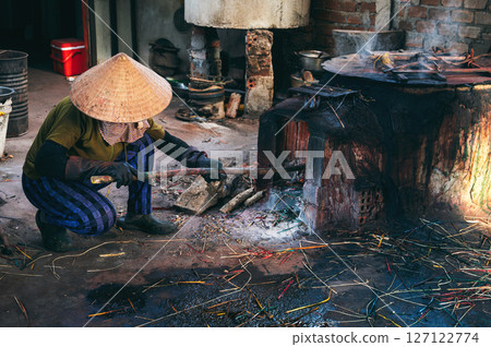 Vietnamese female stoker worker works in straw hat in a workshop and lights a coal furnace at a reed dyeing plant in Vietnam. Woman does dirty hard men's work in a factory in Asia 127122774