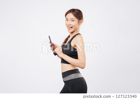Phone in hand and another phone swipe later, this fit sporty woman stands confidently in workout clothes, showing off her gym dedication and mobile lifestyle. isolated studio white background Phone in hand and another phone swipe later, this fit sporty woman stands confidently in workout clothes, showing off her gym dedication and mobile lifestyle. isolated studio white background 127123340