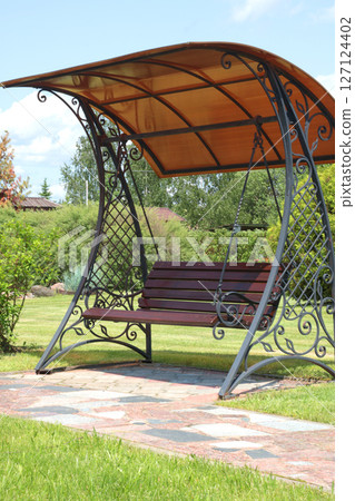 Wrought iron swing with canopy near tile path in park. Green lawn, bushes and trees in summer on sunny day. Background for design. Ecology and nature. Landscape design. 127124402
