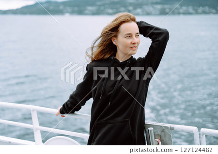 Adult woman with earplugs on ferry in Norway, black hoodie, windblown hair, scenic fjord view 127124482