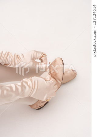 Closeup of woman in satin gloves tying bow ballet flats in balletcore fashion studio shoot. Closeup of woman in satin gloves tying bow ballet flats in balletcore fashion studio shoot. 127124514