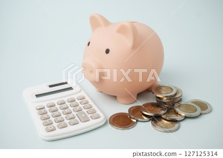 Close up pink piggy bank on light blue background overflowing coins and calculator. 127125314