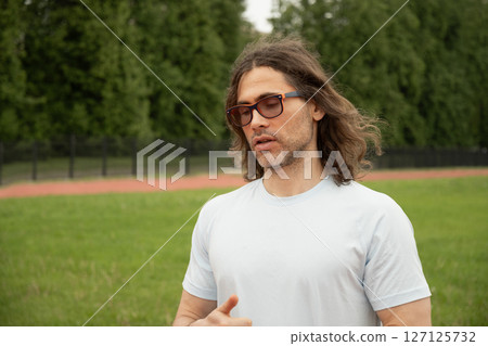 Man or personal trainer with long hair and sunglasses speaking outdoors on sports field. He explains exercises, appears relaxed and thoughtful, wearing white T-shirt against background of green grass Man or personal trainer with long hair and sunglasses speaking outdoors on sports field. He explains exercises, appears relaxed and thoughtful, wearing white T-shirt against background of green grass 127125732