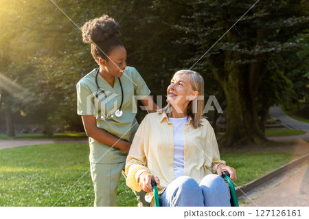 African american nurse rolling a wheelchair with a female patient in a park 127126161