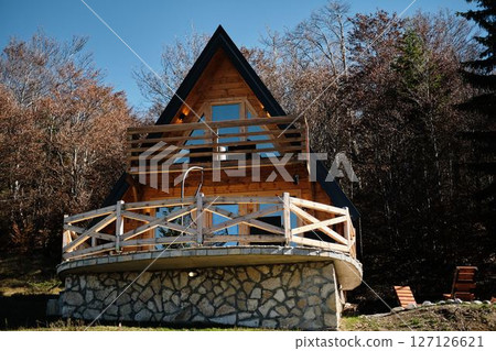 A modern wooden A-frame cabin with a stone foundation stands under a clear blue sky, surrounded by autumn forest in the Montenegro mountains, offering a cozy retreat in nature A modern wooden A-frame cabin with a stone foundation stands under a clear blue sky, surrounded by autumn forest in the Montenegro mountains, offering a cozy retreat in nature 127126621