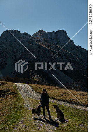 Woman hiking with two dogs - German and Australian shepherds on a mountain trail in the Komovi range, Montenegro, surrounded by dramatic peaks and golden autumn meadows. Hiking with pet concept Woman hiking with two dogs - German and Australian shepherds on a mountain trail in the Komovi range, Montenegro, surrounded by dramatic peaks and golden autumn meadows. Hiking with pet concept 127126629