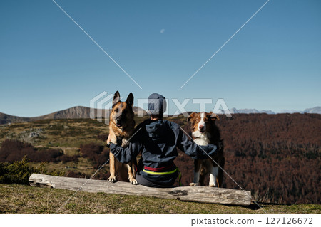 Woman sits on a log with two dogs. German and Australian Shepherds, enjoying peaceful autumn day in Montenegro Komovi mountains with owner. The moment captures loyalty and serenity. Rear view Woman sits on a log with two dogs. German and Australian Shepherds, enjoying peaceful autumn day in Montenegro Komovi mountains with owner. The moment captures loyalty and serenity. Rear view 127126672