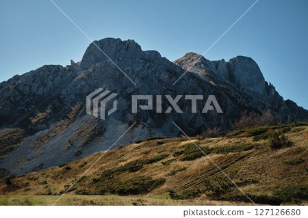 Sharp rocky peaks of the Komovi mountains in Montenegro rise above green autumn shrubs under a clear blue sky. A peaceful scene of natural wilderness and crisp mountain air. Sharp rocky peaks of the Komovi mountains in Montenegro rise above green autumn shrubs under a clear blue sky. A peaceful scene of natural wilderness and crisp mountain air. 127126680