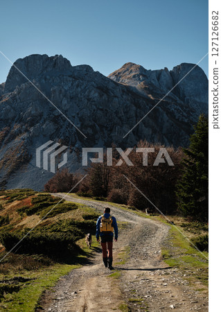 Male solo hiker with a yellow backpack walks along a winding dirt trail in the Komovi mountains in Montenegro, accompanied by two dogs on a sunny autumn day. Rear view. Hiking with pet concept 127126682