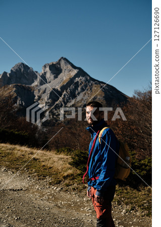 Young male hiker with a backpack stands on a mountain trail in the Komovi range, Montenegro. Dressed in blue and red hiking gear, he enjoys the autumn scenery under a clear sky 127126690