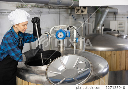 Focused female brewer mixing raw materials in fermenter 127128048