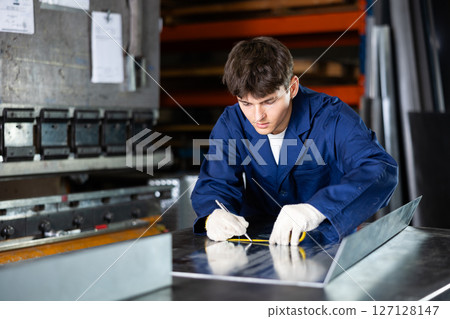 Young worker marking steel sheet with tape measure in workshop 127128147