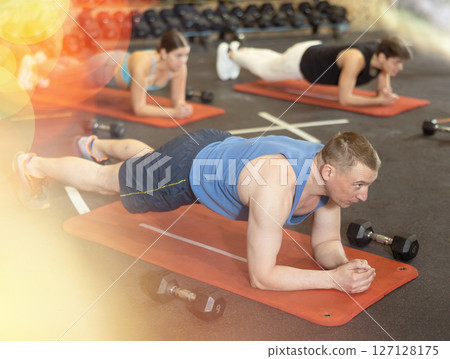 Energetic man doing side-plank near other people in gym 127128175