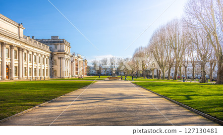 Visitors stroll through the well-maintained gardens of the Old Royal Naval College in Greenwich, enjoying the sunny day and historic architecture framed by leafless trees. 127130481