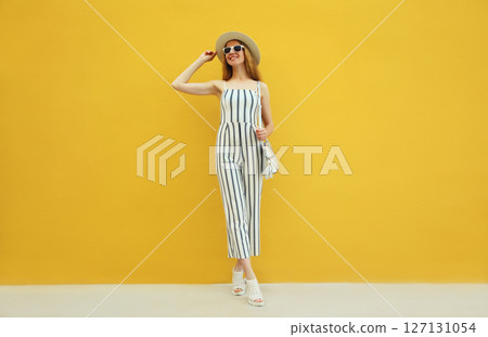 Summer outfit, stylish beautiful young woman posing in white striped jumpsuit against yellow wall 127131054