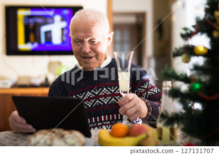 Elderly man wishes Merry Christmas by laptop to friends and acquaintances sitting at the festive table in house Elderly man wishes Merry Christmas by laptop to friends and acquaintances sitting at the festive table in house 127131707