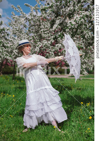 Mature woman of 60 years old in retro style white canotier hat, dress, gloves, glasses and umbrella in blossoming white and pink apple orchard on summer day 127131917