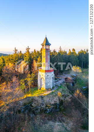 The Stepanka Lookout Tower stands tall against a clear spring sky in Korenov, surrounded by lush greenery. Sunlight illuminates the tower, highlighting its architectural beauty. 127132100