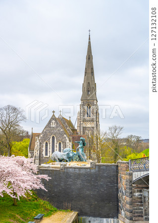 Visitors admire St Alban's Church near Kastellet in Copenhagen, Denmark. The church stands out with its tall spire and beautiful architecture amidst spring blossoms and lush greenery. 127132108