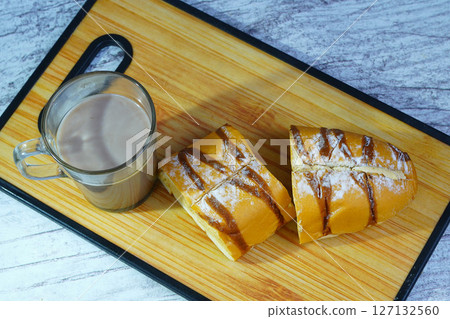 A sliced mocha golden loaf with chocolate drizzle and powdered sugar, served with a glass of hot chocolate on a wooden board. 127132560
