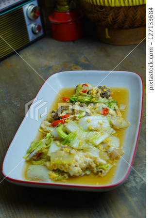 A close-up of a stir-fried dish with napa cabbage, eggs, and meat, served in a white rectangular plate with a red rim, on a wooden table. A close-up of a stir-fried dish with napa cabbage, eggs, and meat, served in a white rectangular plate with a red rim, on a wooden table. 127133684