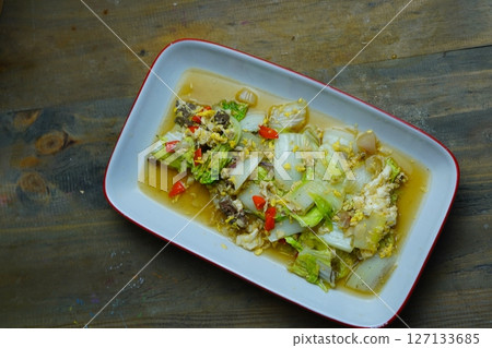 A close-up of a stir-fried dish with napa cabbage, eggs, and meat, served in a white rectangular plate with a red rim, on a wooden table. A close-up of a stir-fried dish with napa cabbage, eggs, and meat, served in a white rectangular plate with a red rim, on a wooden table. 127133685