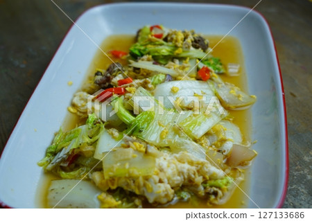 A close-up of a stir-fried dish with napa cabbage, eggs, and meat, served in a white rectangular plate with a red rim, on a wooden table. A close-up of a stir-fried dish with napa cabbage, eggs, and meat, served in a white rectangular plate with a red rim, on a wooden table. 127133686