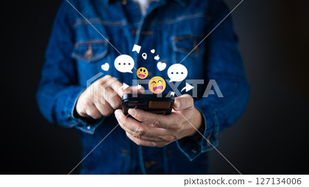 A person in a denim jacket interacts with a smartphone, surrounded by floating emojis, chat bubbles, hearts, and icons representing social engagement and network connectivity in the digital world. A person in a denim jacket interacts with a smartphone, surrounded by floating emojis, chat bubbles, hearts, and icons representing social engagement and network connectivity in the digital world. 127134006