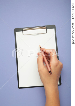 A female hand hold a pen and black clipboard with paper on purple background. Vertical photo 127134320