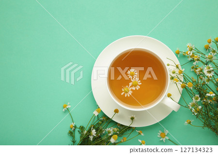 Aromatic herbal tea in white cup and chamomiles on mint background. Space for text 127134323