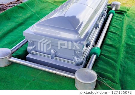 Casket prepared for burial sits on vibrant green turf in calm cemetery ceremonial under bright daylight. 127135072