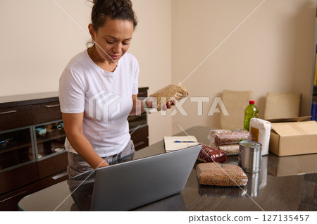 Woman Organizing Food Supplies on Laptop at Home in Kitchen 127135457