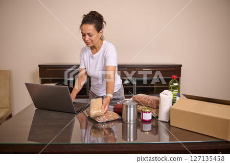 Woman Organizing Pantry Supplies Using Laptop for Inventory and Meal Planning 127135458