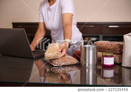 Woman Organizing Non-Perishable Food Items at Home with Laptop on Glass Table 127135459