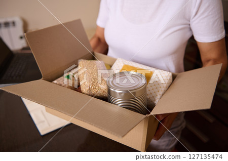 Person Holding A Cardboard Box Filled with Canned Goods and Dry Food Items 127135474