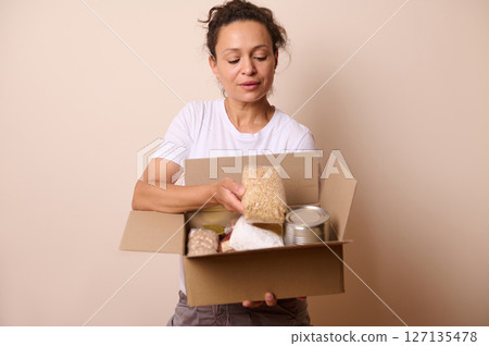 Woman Holding a Box of Canned Food and Grains in a Simple Room Woman Holding a Box of Canned Food and Grains in a Simple Room 127135478
