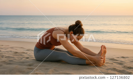 Young woman practicing yoga on sandy beach at sunrise, stretching her legs while seated near the ocean. Calm and focused morning meditation. Peaceful lifestyle concept. 127136802