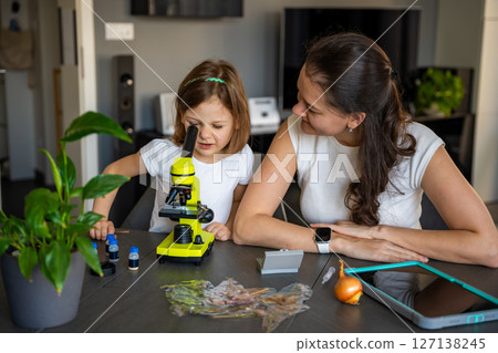 Little girl and her mother examining onion skin under a microscope at home. Concept of family science education and early biology learning 127138245
